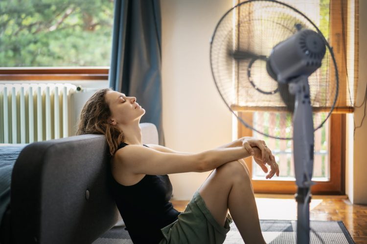 Young woman suffering from heat sitting at home on the floor in front of a fan